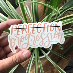 May include: A sticker with the text "PERFECTION progression" in a peachy pink color with a black line through the word "perfection".