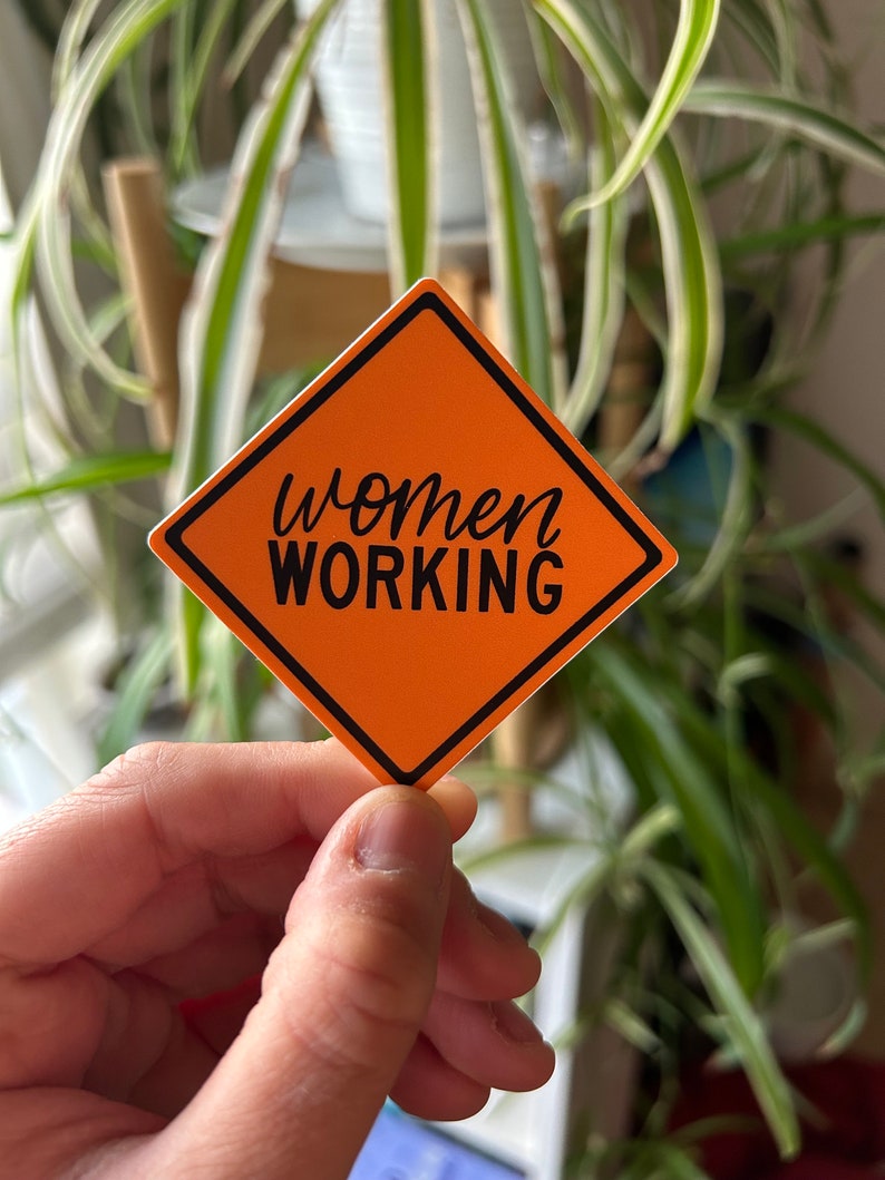 Op de afbeelding: Oranje sticker met een bouwteken met de tekst "women working" in zwarte letters.