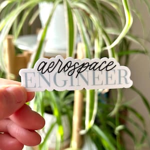 May include: A white sticker with the text "aerospace ENGINEER" in black script. The sticker is being held by a hand against a blurred background of a green plant.