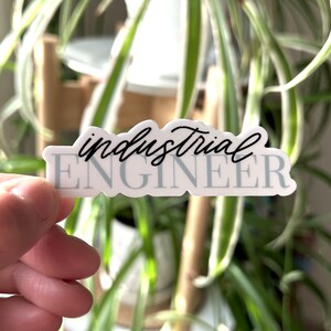 May include: White sticker with the text "industrial ENGINEER" in black script font. The sticker is being held by a hand against a blurred background of a green plant.