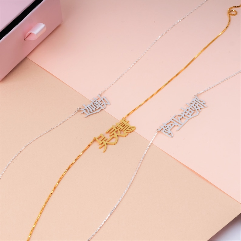 Peut inclure: Trois colliers en argent et en or avec des caract&egrave;res chinois suspendus. Les colliers sont sur un fond rose et beige.