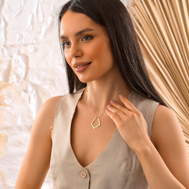 May include: A woman wearing a beige sleeveless top with a gold necklace featuring a geometric pendant. The pendant is shaped like a diamond and has a small inscription inside.