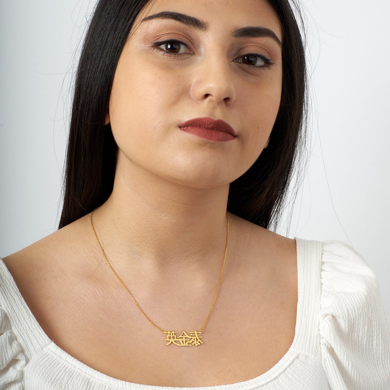 Peut inclure: Un collier en or avec une cha&icirc;ne d&eacute;licate et un pendentif repr&eacute;sentant les caract&egrave;res chinois pour "beau" dans une police moderne et audacieuse.