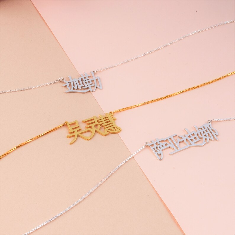 Peut inclure: Trois colliers en argent avec des caract&egrave;res chinois suspendus. Les colliers sont sur un fond rose clair et beige.