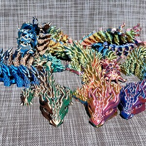 May include: A collection of colorful, articulated dragon figurines in various sizes. The dragons feature a metallic, iridescent finish with hues of blue, green, gold, and red. The detailed designs include spiky spines and textured scales, creating a fantasy theme.