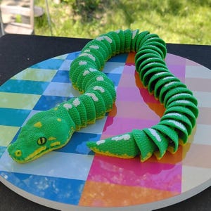 May include: A green and yellow articulated snake toy with white spots. The snake is coiled on a colorful, patterned surface. The snake's head is detailed with yellow accents around the eyes and mouth.