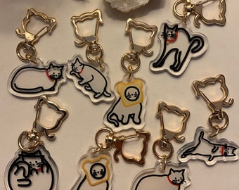 May include: A set of 10 clear acrylic keychains featuring different cartoon cats. The cats are in various poses, including sitting, standing, and lying down. Some of the cats are wearing hats or other accessories. The keychains are attached to gold-colored metal rings.