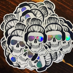 May include: A collection of colorful skull stickers with a bun hairstyle and iridescent sunglasses. The stickers are designed with a black and white outline and feature a variety of colors, including pink, blue, green, and purple.