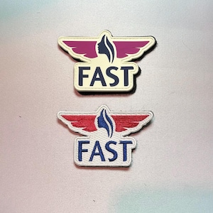 May include: Two embroidered patches with a winged woman design and the word "FAST". One patch features a purple and cream color scheme, while the other has red and blue accents. The patches are on a light pink background.