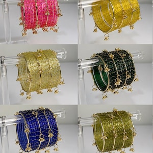 May include: A selection of colourful bangles in shades of pink, yellow, gold, green, blue, and orange. Each bangle features small, dangling charms. The bangles are displayed on clear stands, highlighting their intricate design and vibrant colours.