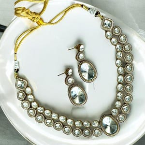 Könnte beinhalten: Ein goldenes Halsketten- und Ohrring-Set mit einer doppelten Reihe von klaren, facettierten Steinen. Die Halskette hat eine zarte goldene Kette und die Ohrringe sind im Tropfen-Stil.