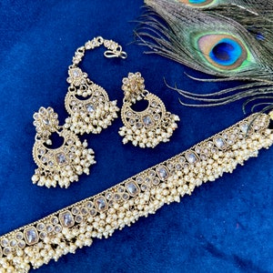 Op de afbeelding: Een gouden Indiase bruidssieradenset met een chokerketting, oorbellen en een maang tikka. De set is versierd met heldere stenen en parel accenten.
