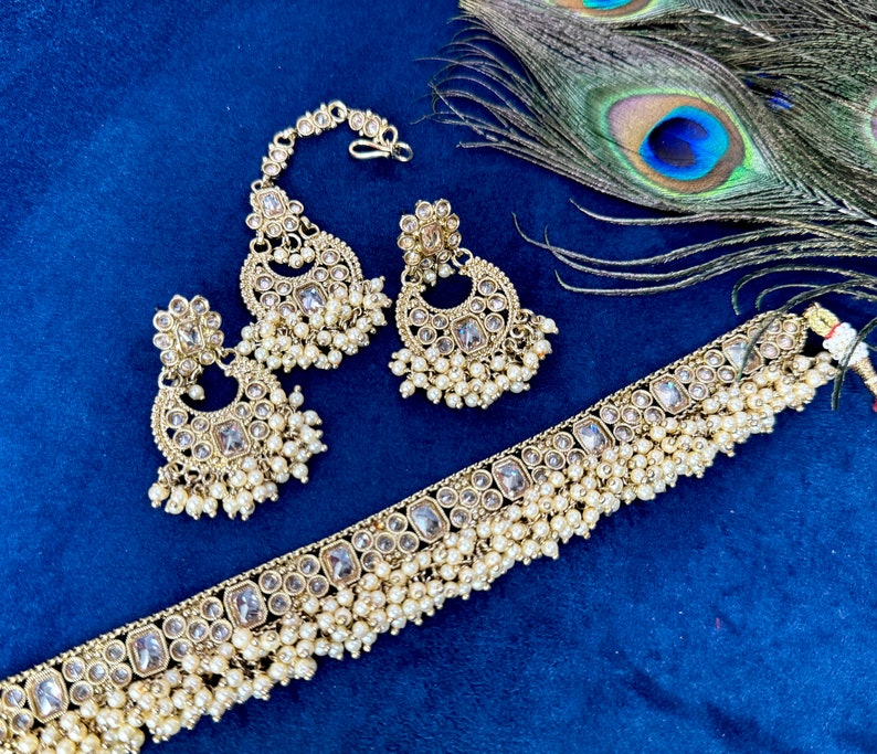 Op de afbeelding: Een gouden Indiase bruidssieradenset met een chokerketting, oorbellen en een maang tikka. De set is versierd met heldere stenen en parel accenten.