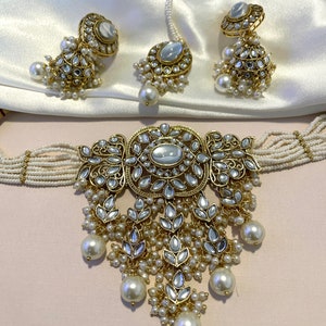 May include: A gold and white Indian bridal jewelry set with a choker necklace, earrings, and a nose ring. The necklace features a large, ornate pendant with a pearl drop and the earrings and nose ring have matching designs.