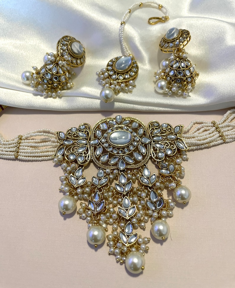 May include: A gold and white Indian bridal jewelry set with a choker necklace, earrings, and a nose ring. The necklace features a large, ornate pendant with a pearl drop and the earrings and nose ring have matching designs.