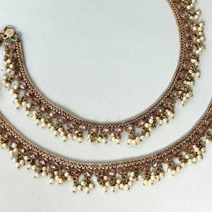Op de afbeelding: Een paar gouden enkelbandjes met roze en witte kralen. De enkelbandjes hebben een delicaat ontwerp met kleine, ronde kralen en een hangende hanger.