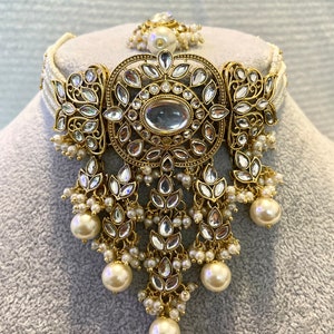 May include: A gold and white pearl choker necklace with a large, ornate pendant featuring a central oval-shaped stone surrounded by smaller stones. The pendant is decorated with intricate floral designs.
