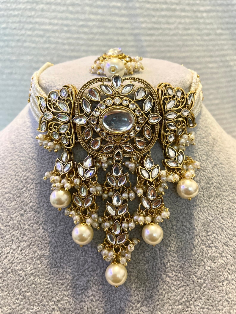 May include: A gold and white pearl choker necklace with a large, ornate pendant featuring a central oval-shaped stone surrounded by smaller stones. The pendant is decorated with intricate floral designs.