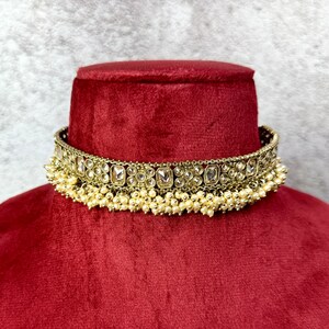 Op de afbeelding: Een gouden choker ketting met een rij heldere stenen en een franje van witte parels.