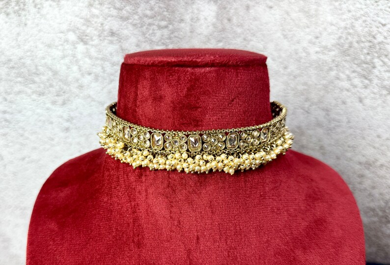 Op de afbeelding: Een gouden choker ketting met een rij heldere stenen en een franje van witte parels.
