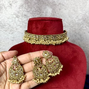 Op de afbeelding: Een gouden chokerketting met heldere stenen en parel accenten. Passende oorbellen met heldere stenen en parel accenten.