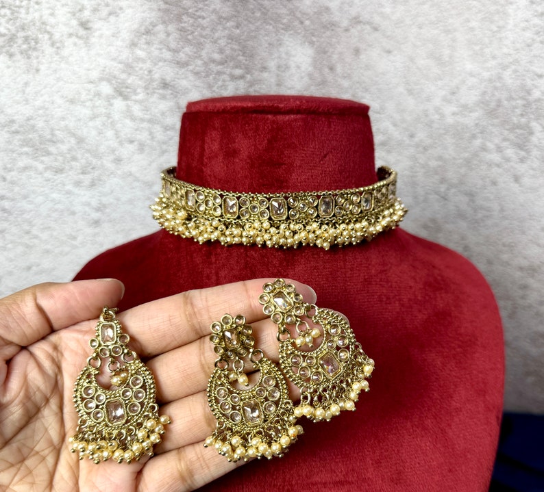Op de afbeelding: Een gouden chokerketting met heldere stenen en parel accenten. Passende oorbellen met heldere stenen en parel accenten.