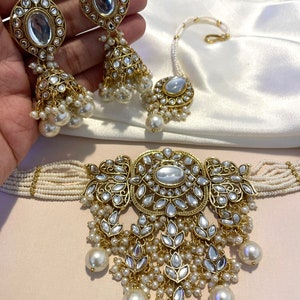 May include: A gold and white Indian bridal jewelry set with a choker necklace, earrings, and a nose ring. The choker necklace is made of white beads and has a large, ornate pendant with a white stone in the center. The earrings are also made of white beads and have a similar pendant to the necklace. The nose ring is a simple gold ring with a white stone in the center.