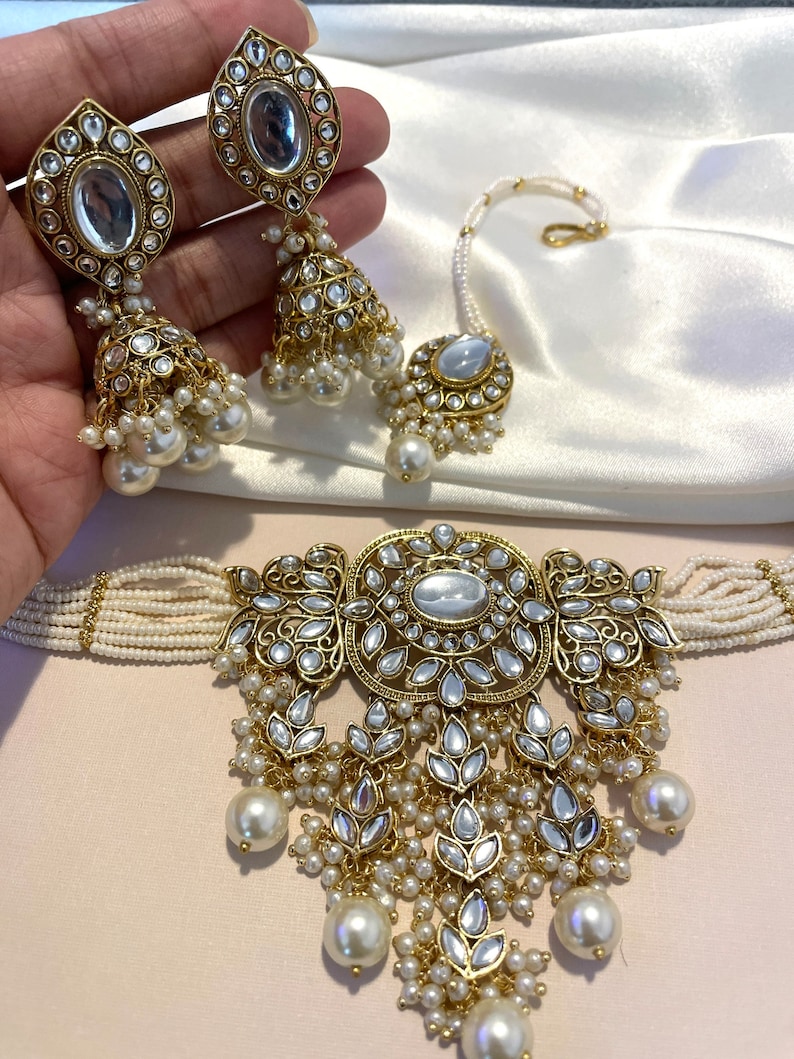 May include: A gold and white Indian bridal jewelry set with a choker necklace, earrings, and a nose ring. The choker necklace is made of white beads and has a large, ornate pendant with a white stone in the center. The earrings are also made of white beads and have a similar pendant to the necklace. The nose ring is a simple gold ring with a white stone in the center.