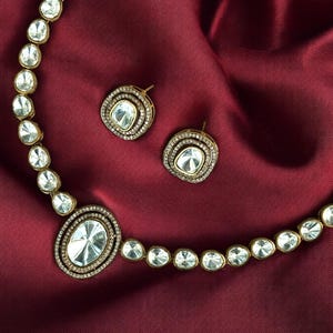 Könnte beinhalten: Eine goldene Halskette mit einem großen ovalen Anhänger und zwei passenden Ohrringen. Die Halskette und die Ohrringe sind mit klaren, facettierten Steinen besetzt, die in Gold gefasst sind.