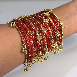 May include: A set of red glass bangles with gold-coloured embellishments and dangling bell charms. The bangles feature floral and twisted rope patterns, giving a festive and ornate look. The jewellery is shown on a wrist.