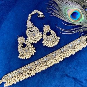 Op de afbeelding: Een gouden sieradenset met heldere stenen en parel accenten. De set bevat een choker ketting, een paar oorbellen en een maang tikka.