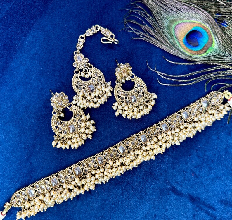 Op de afbeelding: Een gouden sieradenset met heldere stenen en parel accenten. De set bevat een choker ketting, een paar oorbellen en een maang tikka.