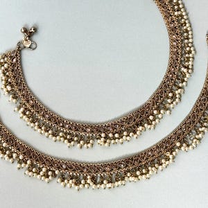 Op de afbeelding: Een paar gouden enkelbandjes met een delicaat ontwerp met kleine, heldere stenen en witte kralen. De enkelbandjes hebben een hangende franje van witte kralen.