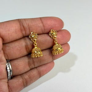 May include: A pair of gold-toned jhumka earrings featuring a flower-shaped stud top connected to a bell-shaped drop. The bell portion displays intricate textured detailing and small dangling beads along the bottom edge of the metal structure.