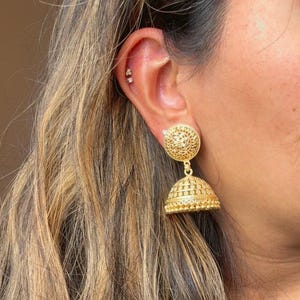 May include: Gold dangle earrings with a round, ornate design. The earrings have a smaller, round, ornate charm hanging from the larger charm.