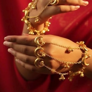 May include: Gold-coloured hand jewellery featuring crescent moon and bell-shaped charms. The jewellery includes rings connected by delicate chains to bracelets. The background is a rich red fabric.