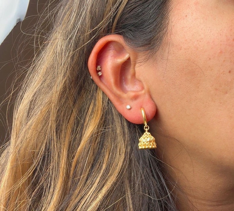May include: Gold hoop earrings with a dangling gold charm in the shape of a bell. The charm has a detailed, intricate design.