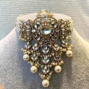May include: A gold and white choker necklace with a large, ornate pendant featuring a round, clear stone surrounded by smaller stones. The pendant is attached to a delicate chain with multiple strands of pearls.