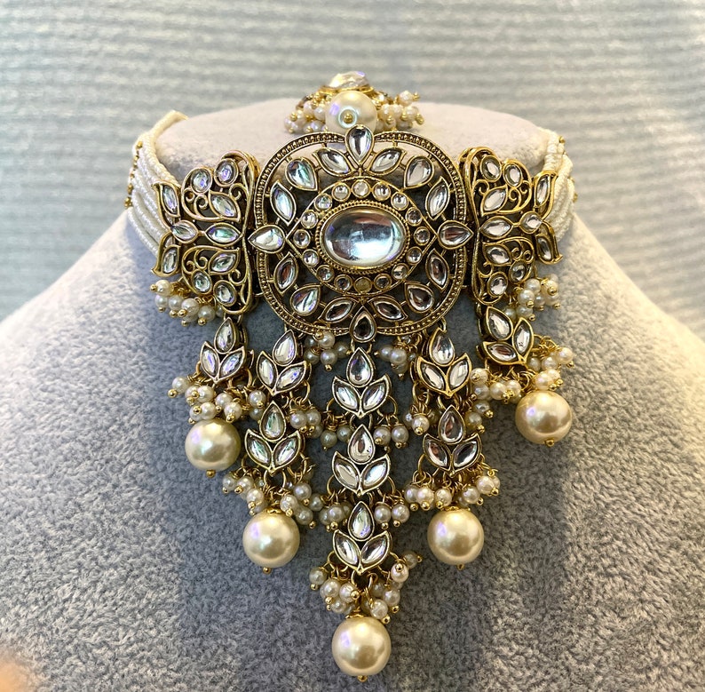 May include: A gold and white choker necklace with a large, ornate pendant featuring a round, clear stone surrounded by smaller stones. The pendant is attached to a delicate chain with multiple strands of pearls.