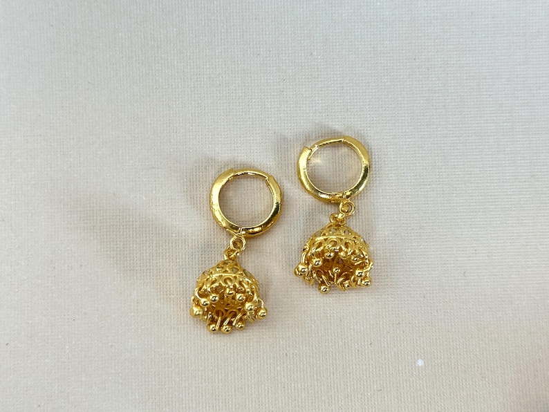 May include: A pair of gold hoop earrings with a dangling bell charm. The bells are intricately designed with a textured surface.