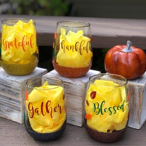 May include: Four clear glass wine glasses with gold, brown, and black glitter bottoms. Each glass has a yellow paper insert with a different word: "grateful", "thankful", "gather", and "blessed". The glasses are decorated with small orange and green leaves.