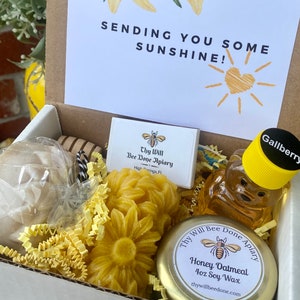 Op de afbeelding: Een geschenkdoos met een stuk zeep in plastic, twee bloemvormige bijenwasstukken, een berenvormige honingfles met een Gallberry-label en een 4oz sojawas kaars. Het deksel toont gele lelies en de tekst SENDING YOU SOME SUNSHINE!