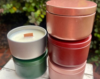 May include: A collection of five round metal candle tins in various colors including copper, dark red, light pink, silver, and dark green. One silver tin is open, revealing white wax and a flat wooden wick. The tins are stacked and arranged together.