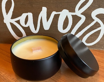 May include: A round black metal candle tin containing off-white wax with a single wooden wick, accompanied by a matching black lid placed to the side. The background features a brown wooden sign with white painted leaf designs and the word favours.