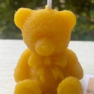 May include: A yellow beeswax candle shaped like a teddy bear, with a white wick. The bear has a bow tie and is sitting upright. The candle is a golden yellow color and has a textured surface.
