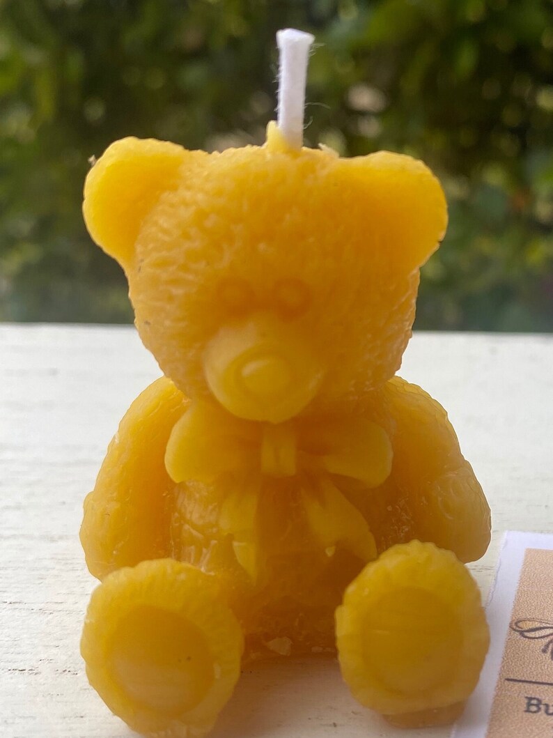 May include: A yellow beeswax candle shaped like a teddy bear, with a white wick. The bear has a bow tie and is sitting upright. The candle is a golden yellow color and has a textured surface.