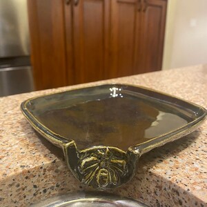 May include: A dark green ceramic dish with a decorative bee design. The dish has a lip on one side that allows it to be hung on the edge of a sink.