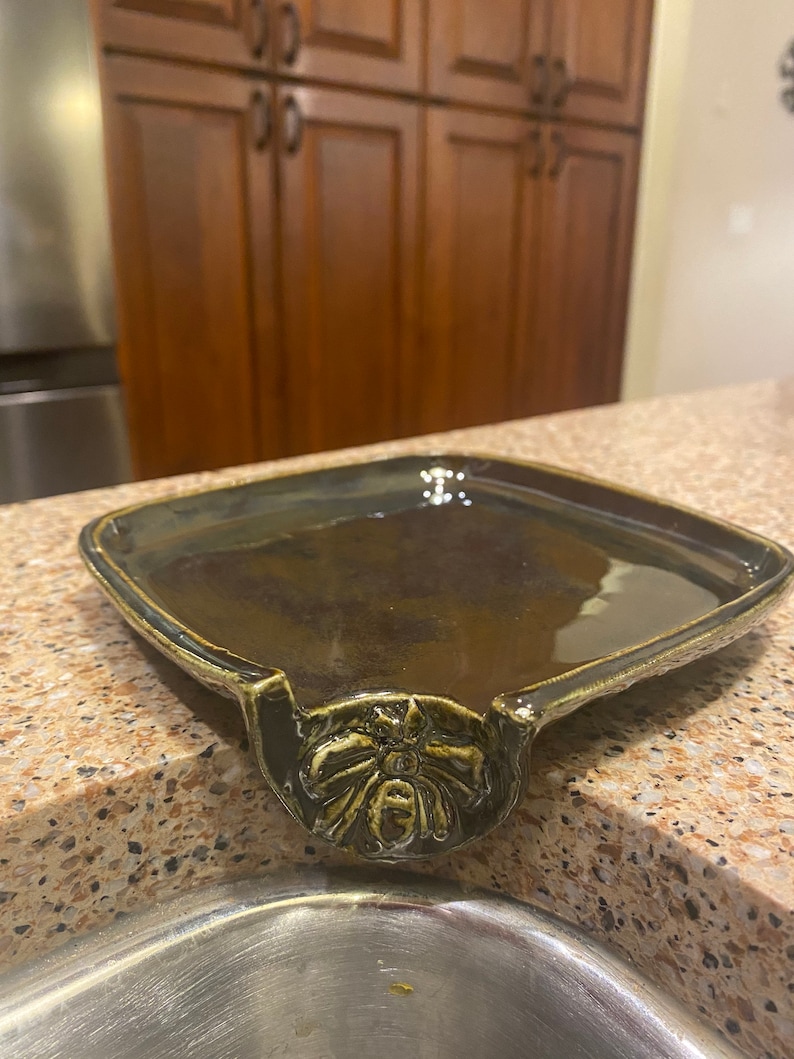 May include: A dark green ceramic dish with a decorative bee design. The dish has a lip on one side that allows it to be hung on the edge of a sink.
