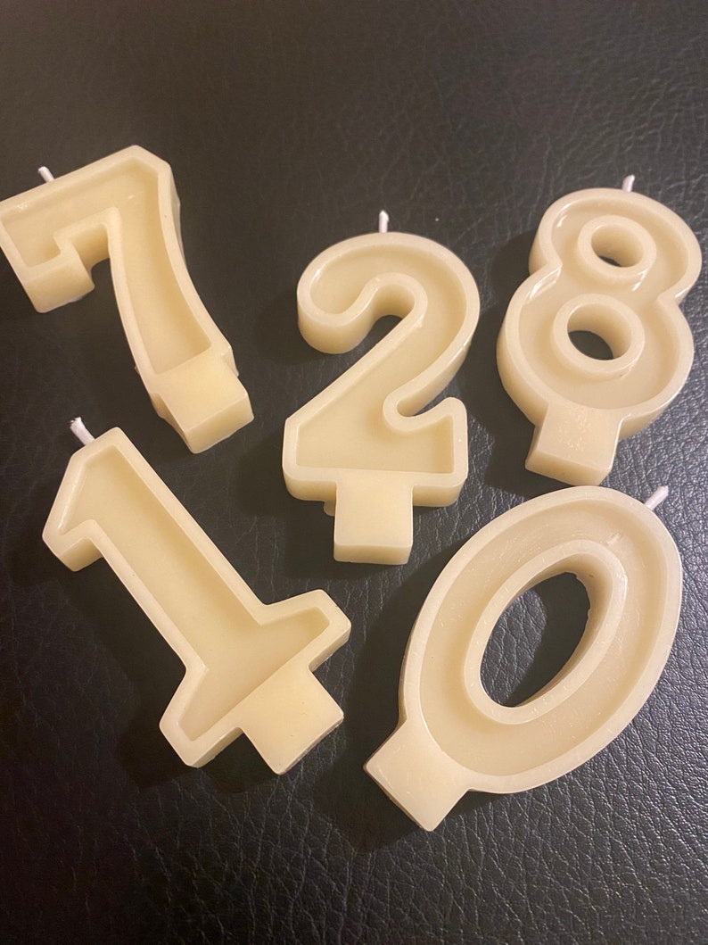 May include: Five ivory-colored number candles: 0, 1, 2, 7, and 8. Each candle has a wick at the top and a flat base. The candles are arranged on a dark surface.