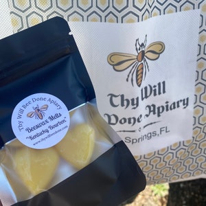 Może przedstawiać: Czarna zamykana torebka zawierająca ż&oacute;łte woski pszczele w kształcie serca z etykietą Thy Will Bee Done Apiary Kentucky Bourbon. Obok znajduje się biała papierowa torba z logo pszczoły i napisem Thy Will Bee Done Apiary Springs, FL.